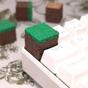 Peut inclure: Un capuchon de clavier Minecraft de couleur verte et marron avec un motif de bloc d'herbe. Le capuchon de clavier est placé sur un clavier blanc avec les touches Tab et Verr. Maj visibles.