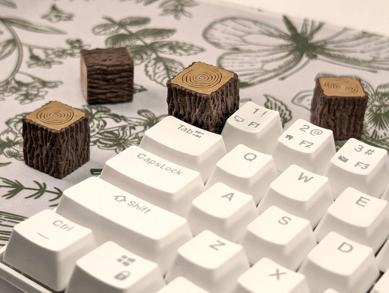 May include: A white keyboard with brown wooden keycaps shaped like tree trunks. The keycaps are on the keys labelled "Tab", "CapsLock", "Shift", "Ctrl", "1! F1", "2@ F2", "3# F3", "Q", "W", "E", "R", "T", "Y", "U", "I", "O", "P", "A", "S", "D", "F", "G", "H", "J", "K", "L", "Z", "X", "C", "V", "B", "N", "M", ".", "/", "Space".
