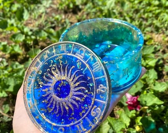 Puede incluir: Una caja de alhajas de resina azul y dorada con un diseño de rueda zodiacal en la tapa. La tapa presenta un sol con rayos dorados y los símbolos del zodíaco alrededor del borde. La caja está llena de purpurina azul y dorada.