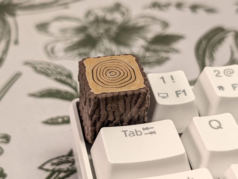 May include: A brown and tan keycap shaped like a tree stump with a wood grain pattern. The keycap is on a white keyboard with the keys labelled "Tab", "1!" and "2@".