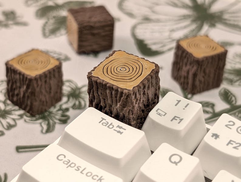 May include: A set of five brown and tan wooden keycaps shaped like tree stumps. The keycaps are arranged on a white keyboard with keys labelled "Tab", "Caps Lock", "1! F1", "Q", and "2@ F2".