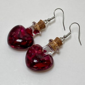 May include: A pair of heart-shaped glass bottle earrings with cork stoppers. The bottles are filled with red rose petals.