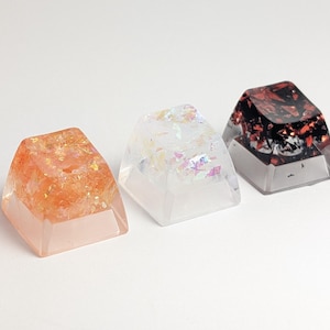 Może przedstawiać: Trzy nasadki klawiszy wykonane z żywicy z różnokolorowym brokatem osadzonym wewnątrz. Nasadki są przezroczyste i mają kwadratowy kształt.