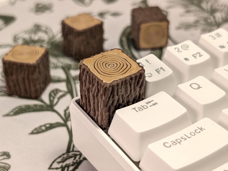 May include: A wooden keycap shaped like a tree stump with a brown and tan colour scheme. The keycap is sitting on a white keyboard with keys labelled "Tab", "CapsLock", "F1", "F2", and "Q".