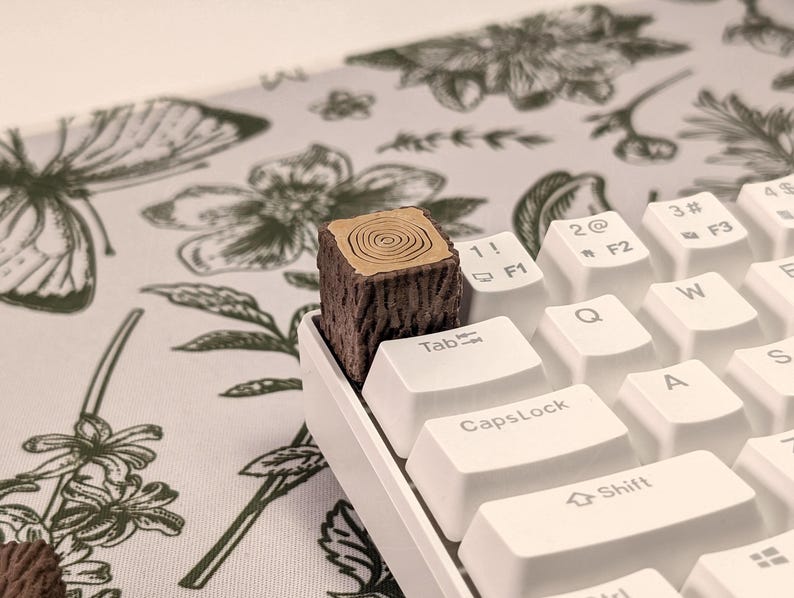 May include: A white mechanical keyboard with a wooden keycap shaped like a tree stump. The keyboard has keys labelled Tab, 1!, 2@, 3#, 4$, F1, F2, F3, Q, W, E, CapsLock, A, S, Shift.