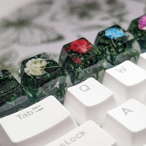 May include: A close-up of a white keyboard with custom keycaps. The keycaps are made of clear resin with embedded flowers and greenery. The keycaps are labeled "Tab", "Q", "W", "A", and "S".