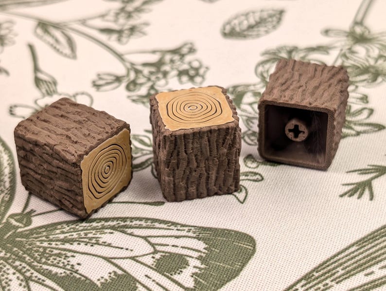 May include: Three brown and tan keycaps shaped like tree stumps. The keycaps have a wood grain texture and a brown ring around the centre of each keycap.