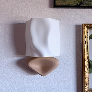 May include: A modern wall-mounted sculpture with a white, textured upper section and a tan, rounded lower section. The sculpture is mounted on a textured white wall, with framed artwork in the background.