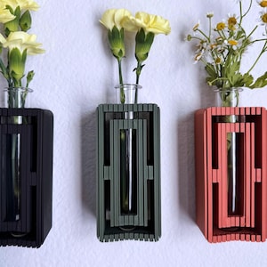 May include: Three wall-mounted vases, each with a glass tube for flowers. The vases are in black, green, and coral, with yellow carnations and white daisies. The vases have a geometric, layered design.