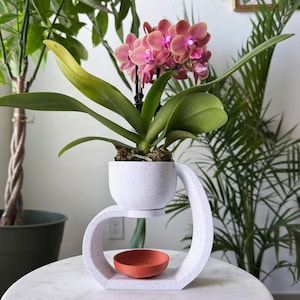 May include: A white ceramic plant stand with a curved design holds a small white pot with a pink orchid plant. The stand has a small, round, orange saucer underneath the pot.