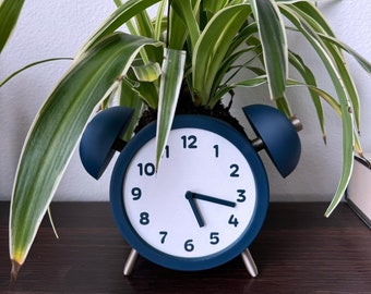 May include: A navy blue alarm clock repurposed as a planter, with a white clock face and black numbers. The clock hands point to 5:20. The clock holds a green and white spider plant, creating a unique decorative piece.