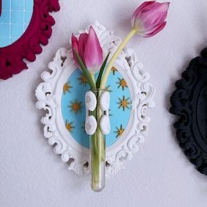May include: A white, ornate wall-mounted vase with a light blue background featuring gold star accents. A clear glass tube holds two pink tulips. Decorative frames in magenta and black are visible.