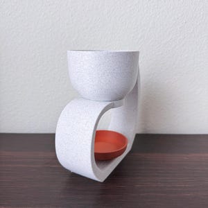 May include: White ceramic wax warmer with a round top and a curved base. The warmer has a small, orange ceramic dish for wax melts.