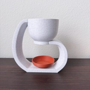 May include: White ceramic diffuser with a curved design. The diffuser has a small, round, orange bowl in the center.
