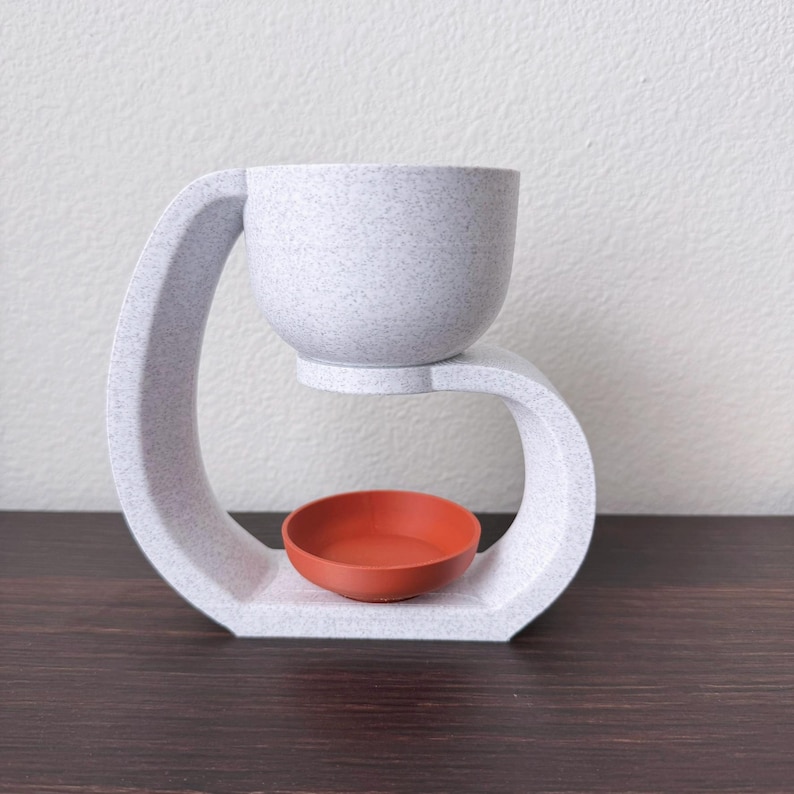 May include: White ceramic diffuser with a curved design. The diffuser has a small, round, orange bowl in the center.