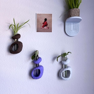 May include: A collection of wall-mounted planters in various colors. The planters hold small plants in glass tubes. One planter is brown, one is purple, and one is light blue. A small painting and a shelf with a plant are also visible.