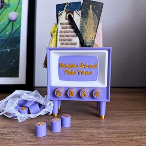 May include: A lilac-coloured TV-shaped book tracker with the text "Books Read This Year" in gold. The tracker has four dials with numbers and gold-tipped legs. Several bookmarks are inside the TV, and small purple cylinders are in front.