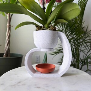 May include: White ceramic plant stand with a curved design. The stand has a small, round, terracotta-colored saucer in the center. A white ceramic pot with a light gray speckled finish sits on top of the stand, holding a green orchid plant with pink flowers.