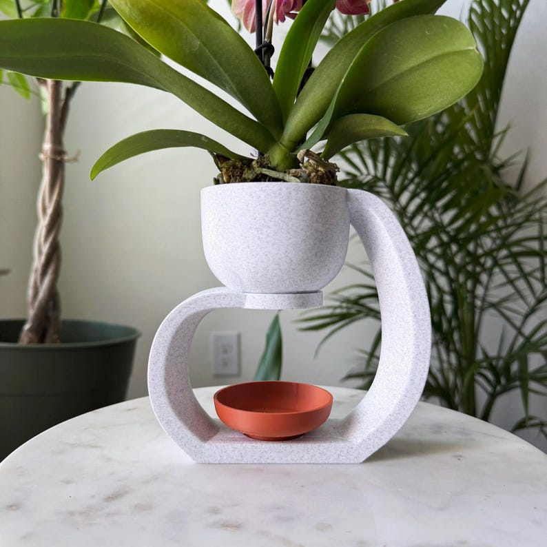 May include: White ceramic plant stand with a curved design. The stand has a small, round, terracotta-colored saucer in the center. A white ceramic pot with a light gray speckled finish sits on top of the stand, holding a green orchid plant with pink flowers.