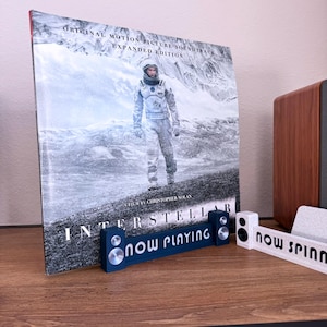 May include: An "Interstellar" vinyl record with a movie still of an astronaut. A blue "now playing" stand and a white "now spinning" stand are in front of the record. The record is on a wooden surface.
