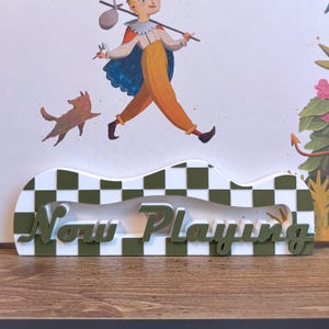 May include: A decorative sign with the words "Now Playing" in green, set against a white and green checkered pattern. The sign is in front of a whimsical illustration of a boy and a dog.