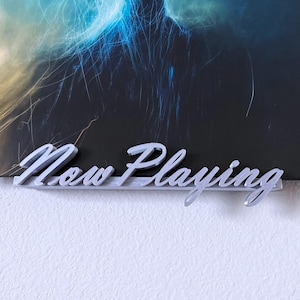 May include: A silver 'Now Playing' sign in a cursive font, positioned below a framed album cover with a dark, abstract design. The sign is mounted on a white wall.