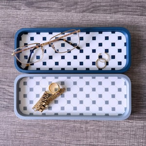 May include: Two rectangular trays with a geometric pattern. The top tray is blue with white squares and the bottom tray is gray with white squares. The top tray has a pair of glasses and two gold rings. The bottom tray has a gold watch band.