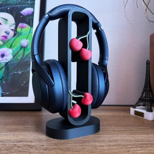 May include: Black headphones on a black headphone stand with three red cherry decorations. The stand has a curved design and a circular base. A framed artwork and a miniature Eiffel Tower are in the background.