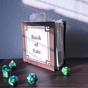 May include: A brown and white decorative book titled "Book of Fate" with a white crown on top. Several green and blue dice are scattered on a dark wooden surface, suggesting a tabletop game theme.