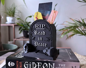 Puede incluir: Un organizador de escritorio negro con forma de lápida con el texto "RIP Read in Peace". El organizador contiene varios artículos, incluyendo un accesorio de cuero amarillo y una tarjeta rosa. Dos pequeñas calaveras están frente al organizador.