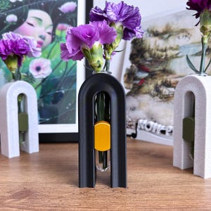 May include: Three modern bud vases, each holding purple carnations. The vases have an arched design, with one black, and two in a speckled white. Each vase has a colored accent piece. The vases are on a wooden surface.