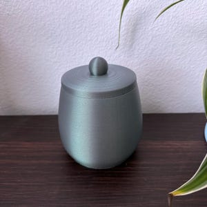 May include: A small, round, grey container with a lid. The container has a smooth, iridescent finish.