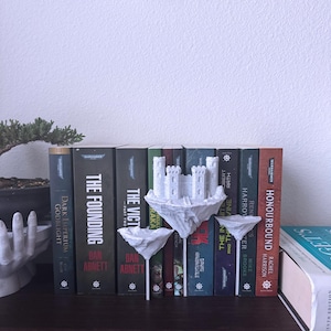 Puede incluir: Separadores de libros blancos impresos en 3D con forma de castillo flotante con torres. Los separadores de libros sostienen una fila de novelas de fantasía con títulos como "The Founding", "The Victory" y "Honourbound".