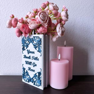Puede incluir: Un jarrón decorativo con forma de libro con detalles florales azules y el texto "Your Book Title Will Go Here". Contiene flores rosas y está junto a dos velas rosas.