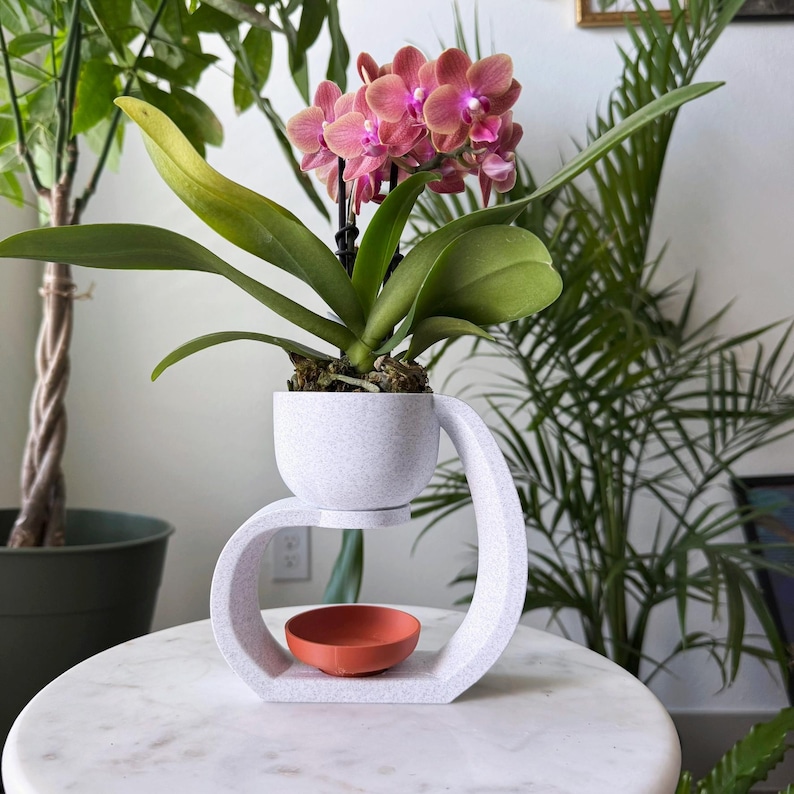 May include: A white ceramic plant stand with a curved design holds a small white pot with a pink orchid plant. The stand has a small, round, orange saucer underneath the pot.