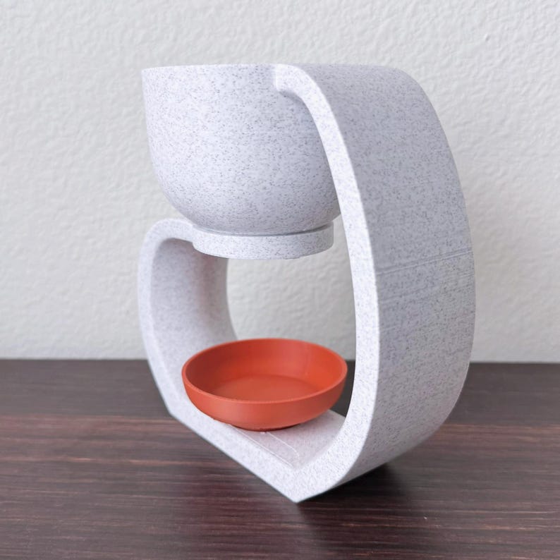 May include: A white ceramic wax warmer with a speckled finish. The warmer has a round bowl on top and a smaller bowl underneath for melting wax.