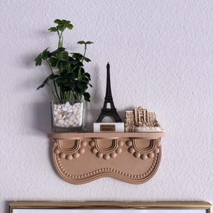 May include: A decorative wall shelf in a light brown hue, adorned with a faux plant in a glass vase, a black Eiffel Tower replica, and a miniature Roman fountain. The shelf has a scalloped design.