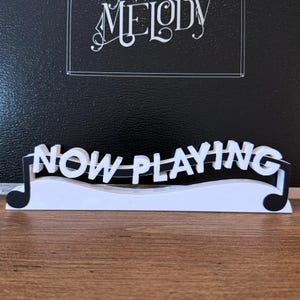 May include: White 3D printed sign with black musical notes and the text "NOW PLAYING" in black letters.