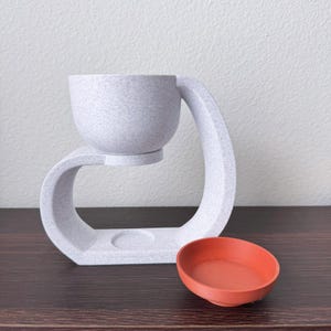 May include: White ceramic diffuser with a round bowl and a curved base. The diffuser is on a dark wood surface with a small orange bowl next to it.