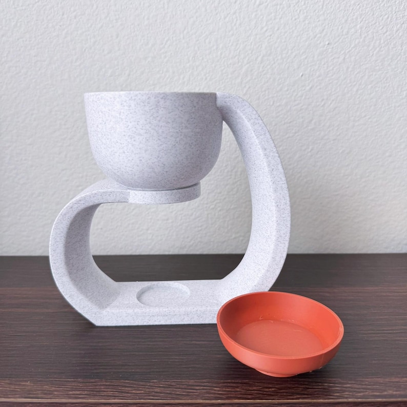 May include: White ceramic diffuser with a round bowl and a curved base. The diffuser is on a dark wood surface with a small orange bowl next to it.