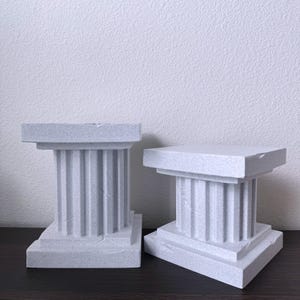 May include: Two white, decorative, fluted, square, pedestal bases. The bases are made of a textured material and are similar in size and design.