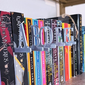 May include: A shelf of colorful books is held in place by silver metal bookends spelling out the word "ABODE". The books have various colored spines and titles. The bookends are in a decorative, cursive font.