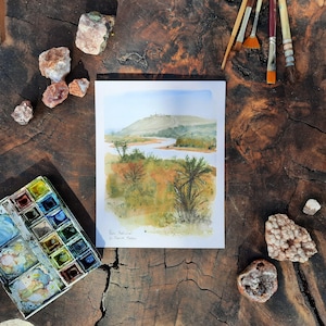 Puede incluir: Acuarela de un paisaje con un río, árboles y colinas. La pintura está rodeada de pinceles, una paleta de acuarela y rocas sobre una superficie de madera. La pintura está firmada "Parc National de Sierra Nevada".