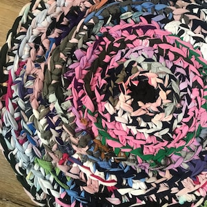 Può includere: Un tappeto rotondo realizzato con strisce di tessuto intrecciate in una tavolozza multicolore che include sfumature di rosa, nero, blu navy, bianco, grigio e verde. Il materiale tessile è intrecciato in un motivo a spirale continua.