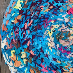 Può includere: Un tappeto circolare realizzato con strisce di tessuto intrecciate in un vivace mix di colori tra cui blu brillante, turchese, magenta, giallo, arancione, marrone e crema. La superficie tessile presenta un motivo denso, strutturato e multicolore.