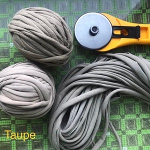 May include: Three balls of taupe colored yarn and a yellow rotary cutter with a black handle. The yarn is made of fabric scraps and is a popular material for crafting.