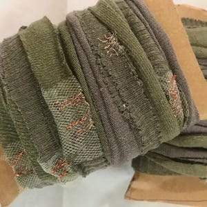 Puede incluir: Carretes de cinta de tela verde oliva y gris con un aspecto desgastado y texturizado. La cinta está adornada con hilo metálico color cobre y una superposición de malla blanca. Los carretes están montados sobre cartón marrón.