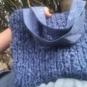 May include: A blue fabric tote bag with a woven texture. The bag has two handles made of a lighter blue fabric, with white stitching and a decorative design. The bag is held by a person.