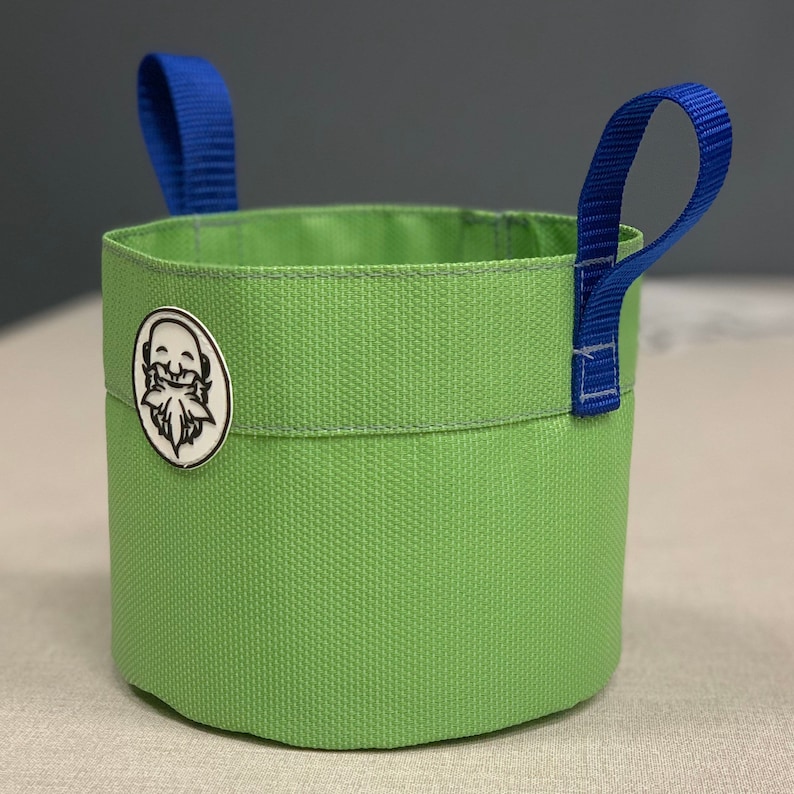 May include: A green fabric bucket with blue handles. The bucket has a white logo of a smiling face with a beard and a leaf.