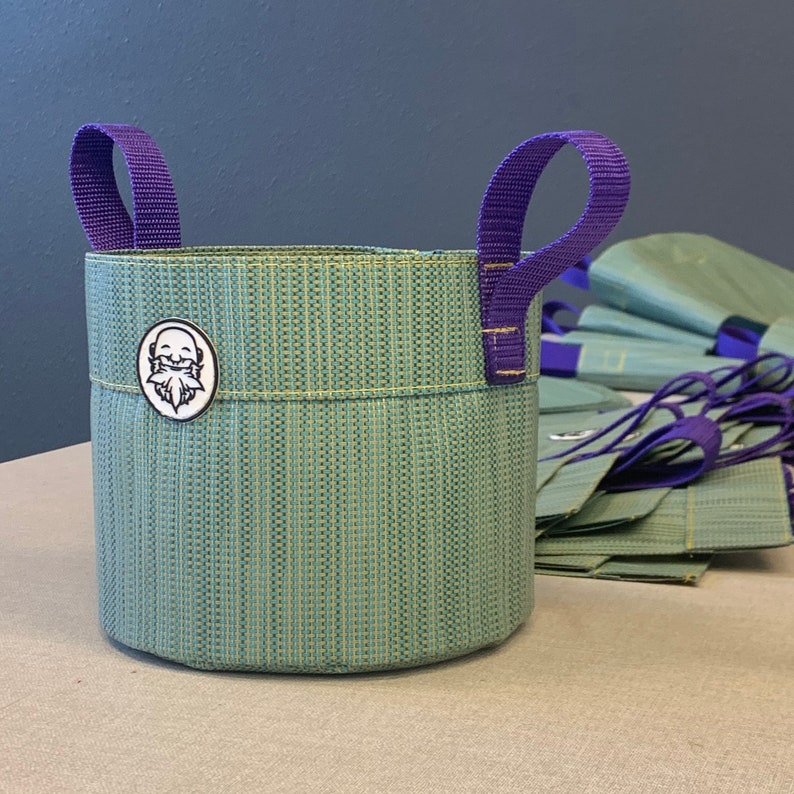 May include: A green fabric planter with purple handles and a white logo of a bearded face. The planter is sitting on a table with a stack of green fabric bags behind it.